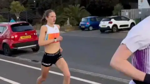Gavin Pavey Innis Fitzgerald is pictured running in Exmouth. She is wearing a white and orange crop top and black shorts. She has brown hair, tied in a ponytail. There are cars parked along the road behind her.