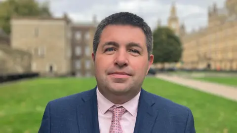 A man is standing in front of Parliament buildings on a green lawn. He is wearing a navy blue suit jacket with light pink shirt and pink patterned tie