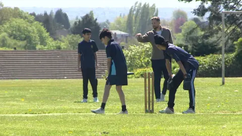 Three young cricketers and Woakes are near the stumps. Trees and bushes are in the background.