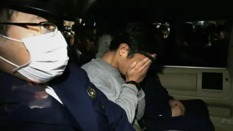 Suspect Takahiro Shiraishi (in grey sweater) covers his face with his hands as he is transported to the prosecutor's office from a police station in Tokyo on 1 November, 2017. He is seated in between police officers in blue uniform.