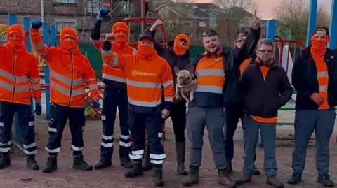 Dale Brown/Pink Mango Socials A group of men wearing high-vis orange workwear and orange balaclavas stand with their fists raised in the air on a playground.