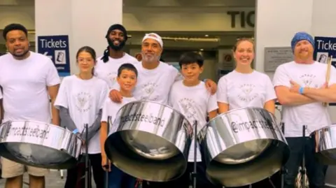 Eight people from the steel band. they are in white t-shirts that say "impact steel band". They are standing in front of three silver steel drums.