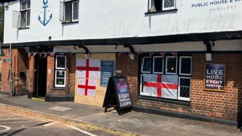 Alexander Stevanovic/BBC The Anchor Inn can be seen with wooden boards covering the damage.
St George's flags are also hanging on it.