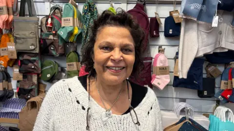 Martin Heath/BBC Caroline Gates with long black hair wearing a white sweater and split glasses on a string. There are clothes and bags on display behind her.