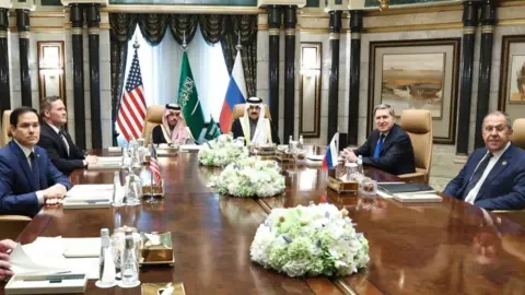 Six men in suits and formal dress sit around a brown table adorned with white and green floral arrangements during a meeting 