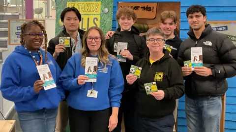 A group of seven people stand in front of a blue painted shed, which is located indoors and next to a staircase. The people in the group are holding leaflets, and posing for the photograph.