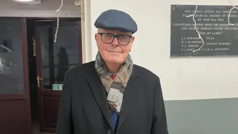 John Fitter wears a grey flatcap, dark rimmed glasses, and has a multi-coloured scarf and black overcoat on. He is standing in front of a doorway with a memorial plaque to his left.