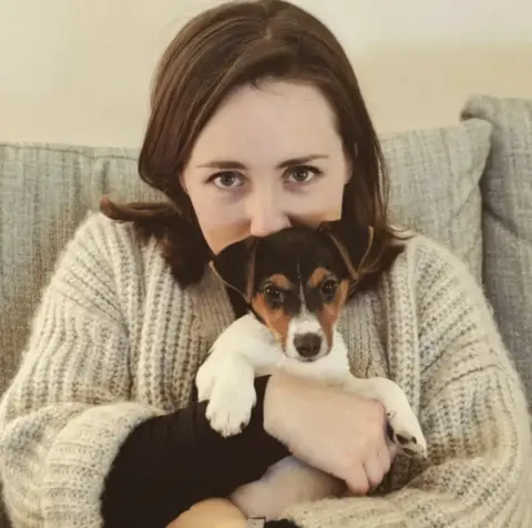 Instagram Person in a cream knitted sweater sitting on a grey sofa, holding a small black, brown, and white puppy close to their chest. The puppy faces the camera with its front paws resting over the person's arm, while a neutral-colored wall and sofa cushions form the background.