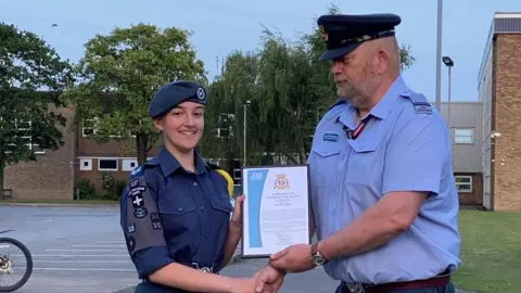 Family handout A teenage girl dressed in a navy RAF cadet uniform and a beret is smiling at the camera whilst holding a framed certificate and shaking the hands of a man dressed in a RAF uniform. They are stood in a car park in front of buildings.