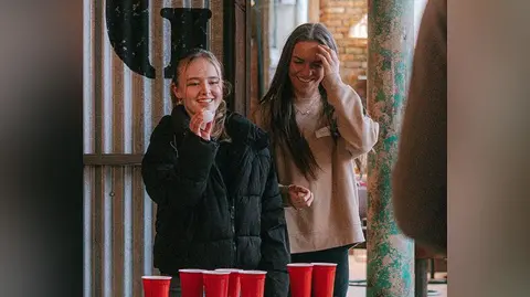 A woman with long blond hair wearing a black coat aims a white ball in a game of beer pong as her friend looks on. They are both laughing.