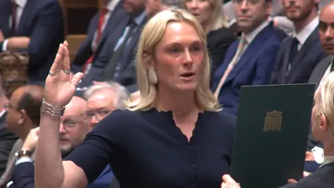 Lara Bird, who has shoulder length blonde hair, stand with her right hand raised as she reads off a green sheet held to her face. 