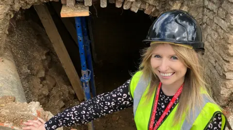 Henry Godfrey-Evans/BBC A woman in a hard hat and a high-vis yellow vest proudly squatting in front of a tunnel entrance. She is smiling.
