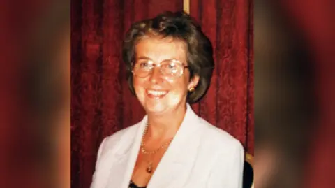 Short Family Veda Short smiles at the camera, she has short brown hair and round silver glasses. She is wearing gold jewellery, a white blazer and black top.