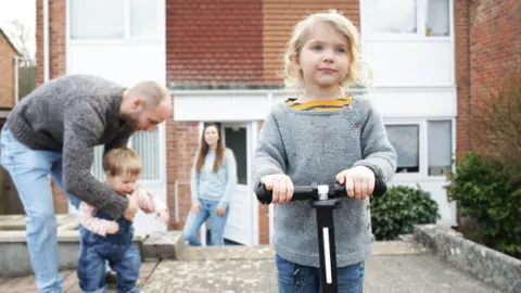 A family are out in front of a house. In the foreground is a young girl with shoulder length blonde hair holding onto the handles of a scooter. In the background a man is helping a toddler to stand and further behind that a women looks towards them.
