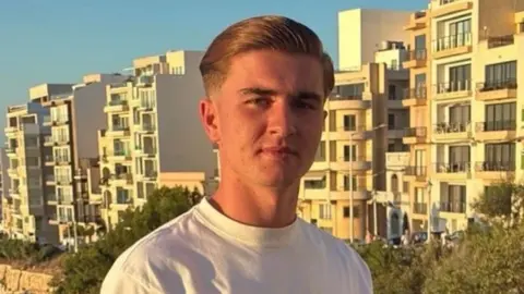 Handout A young with shirt blonde hair wearing a white t-shirt. He is stood in front of a number of apartment blocks, lit up by evening sun. He is looking into the camera with no expression on his face.