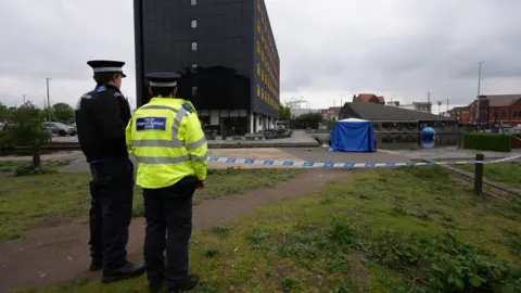 Two police officers stand with their backs to us by blue and white police tap with surrounds paths, grass and paving - with a blue tent on the paving.