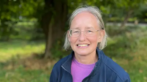Dr Mitchell is a woman with white hair and glasses in a purple T-shirt and blue jacket. She is smiling against a backdrop of trees.
