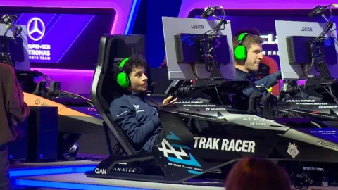 Young man with bright green headphones sitting in gaming chair in front of a screen driving a gaming wheel