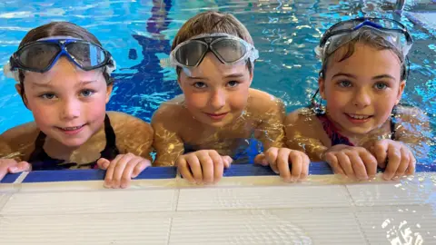 BBC/Lucinda Adam A boy and two girls aged 8 look up from the side of a swimming pool. They are submerged to their shoulders and all three have goggles on their heads.