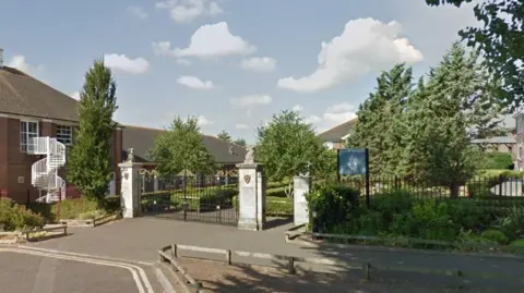 Google Street View of Thomas Hardye School in Dorchester. The two-storey brick buildings are mostly obscured by trees but the entrance is made up of stone pillars and wrought iron gates and railings.