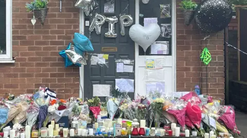 PA Media Candles, floral tributes and balloons outside a house. There are silver balloons spelling out the letters "RIP" hanging across the house's front door.