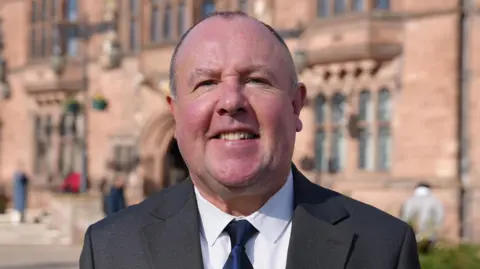 Jim O'Boyle smiles at the camera in front of Coventry's Council House. He is wearing a grey suit jacket and a white shirt with a navy blue tie. 