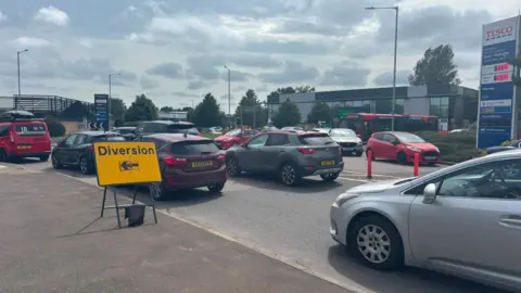 Jaimielee Rendall/BBC Standstill traffic on the approach to a roundabout. On the left of the image is a yellow Diversion sign with an arrow pointing left. In the background is a Pets at Home and a Tesco sign.