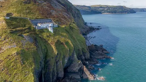 Shaun Davey Foreland Point Lighthouse in Exmoor National Park. It is a large white building with a stone wall behind it, separating it from the moorland. In front of it is the white lighthouse with a wraparound balcony. It is situated very close to the cliff edge, where below, waves break around the jagged rocks. The water is a bright blue and in the background there is another headland stretching out into the sea.