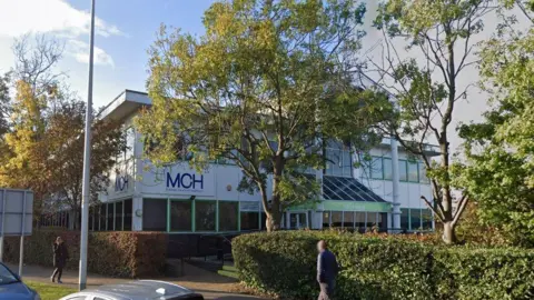 The outside of a two storey building with dark windows and white cladding. There is a glass porch and the letters MCH on a sign on the side of the building. In the foreground are trees and hedges in front of the building.