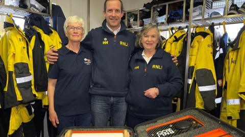 A white woman with white hair, a middle-aged man, and a middle-aged woman, all dressed in RNLI-branded clothing, stand in front of an orange box containing the RNLI scroll. The man as his arms around the women and they are smiling. In the background are a series of yellow waterproofs.