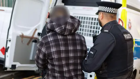 GMP A police officer in uniform holds the arm of a man in a dark purple and white chequered hoodie as he guides him to the back of a police van. The suspect's face is blurred.