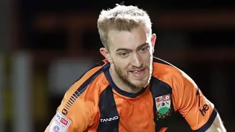 Callum Stead runs after the ball in his amber and black Barnet home shirt. He is looking down at the ball which is out of the picture.