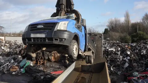 A blue flatbed truck is suspended in the air, being held by a grabber. In the background you can see a scrapyard with lots of metal