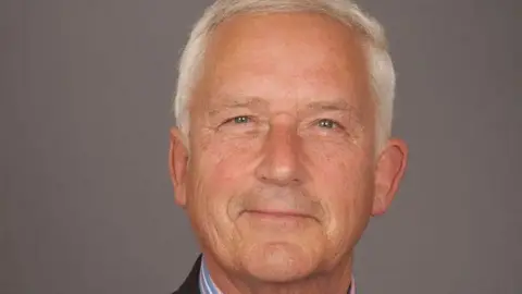 Glen Sanderson has white hair and is looking at the camera. He is wearing a suit and striped shirt. 