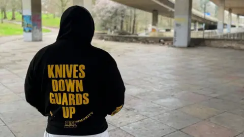 Shelim Ali Shelim Ali. He has his back turned to the camera. He is wearing a black hoodie with the hood up. It has "Knives Down Guards Up" written on the back in big yellow letters.