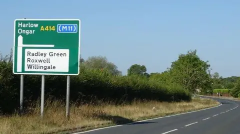A large green road sign telling motorists to continue forwards for Harlow and Ongar, or turn left to Radley Green, Roxwell and Willingale. The road has long grass and bushes next to it.
