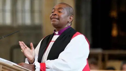 Rose Hudson-Wilkin speaking in front of a microphone on a stand while wearing a white shirt, a red and black  waistcoat and pear earrings. 