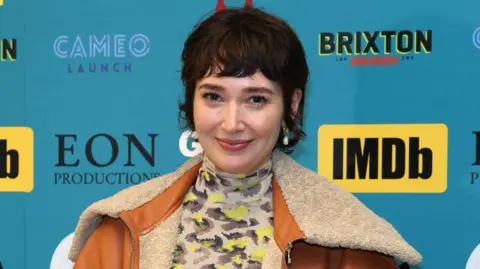 A woman looking at the camera and smiling, she has dark hair and is wearing a brown coat with cream fur. She also has a top with a yellow and brown design which covers her neck. She is standing in front of a board which has sponsor logos including 'IMDb', 'Brixton Brewery', 'Cameo Launch' and 'EON Productions'.