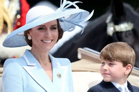 Getty Images Princess Catherine smiles while sitting next to Prince Louis in a carriage, She is wearing a light blue wide-brimmed hat and a light blue jacket with white-trimmed lapels and a brooch. Louis is wearing a suit with a light blue tie.
