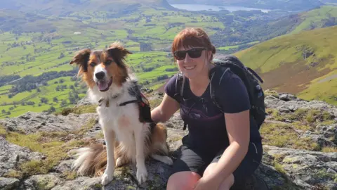 Sam Naylor sits on a rock formation at the top of a fell or mountain with lots of green land behind her - she is wearing a purple t-shirt and black shorts, sunglasses and a backpack. Her dog is a ginger, brown and white collie and has his mouth open and is looking at the camera.