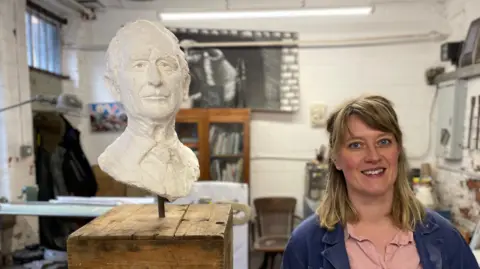 Mark Ansell / BBC Sculptor Lily Marsh stands next to a white plaster version of her King Charles bust in her studio.