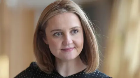 A woman with shoulder-length blonde hair, looking off camera. She is wearing a black and white polka dot top.