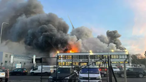 Large plumes of smoke are coming from behind the building at the front of the image. It has a sign that reads 'Fusco Vehicle Sales'. Orange flames are rising to just above the height of the building. Cars are parked in front car park. 