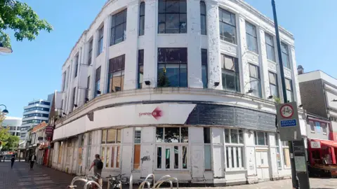 The Poirot building in Swindon town centre. It is a white art deco building on the corner of the street. It is boarded up, with paint flaking off its exterior. A cycle rack is outside on the pavement.