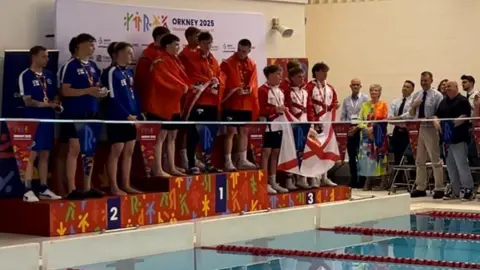 BBC The podium for the men's 4x50m freestyle relay, with Manx swimmers Zachary Bellhouse, Magnus Kelly, Harry Robinson and Joel Watterson in the middle draped in red Manx flags. They are flanked by teams from the Faroe Islands in blue in second, and Jersey in white and red in third. The podium is poolside and there are a number of officials looking on from the right.