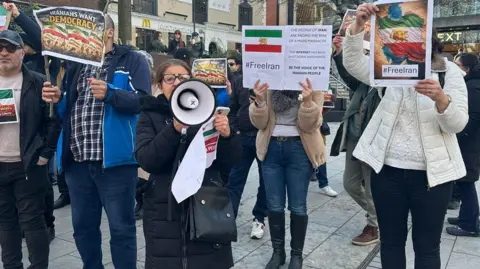 Protesters on a city centre street in Norwich. In the foreground, a woman wearing glasses and a dark coat is speaking into a loud hailer. She is holding a poster and her handbag. Behind her, other protesters are holding up posters saying "Free Iran" and "Iranians Want Democracy".