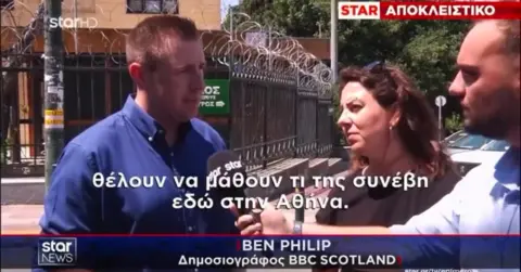 Television news screenshot showing an outdoor interview beside a metal security fence topped with razor wire. Three people are visible in the foreground, with one person speaking into a microphone while another holds recording equipment. Greek-language captions appear across the screen, along with a news banner and the text "STAR ΑΠΟΚΛΕΙΣΤΙΚΟ" at the top. Trees, fencing, and a building are visible in the background.