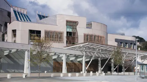 The front of the holyrood building on a cloudy day, with white stone walls wooden slatted windows and small trees.