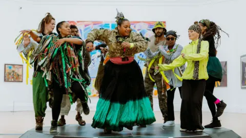 RWA A group of eight dancers mid-movement. A woman centre of the group is wearing a khaki long sleeved top with a black and green long skirt. Other dancers are wearing a mix of outfits, with one woman in a lime green top and black trousers and others with green and brown long ribbon-style material over their shoulders. They are wearing boots and trainers and all look like they are enjoying the dance.