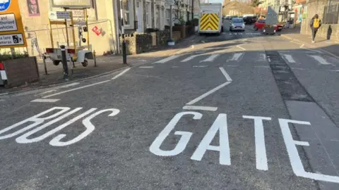 Claire Cavanagh The picture shows the words "bus gate" painted on the road in white on the grey tarmac. There are buildings and vehicles in the background.